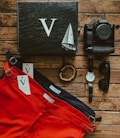 A collection of items laid out on a wooden surface. The items include three pairs of pants in red, navy, and black from the brand Cove Five, a black box with a large white letter V and a sailboat illustration, a wooden bead bracelet, a black watch with a white face, black sunglasses, and a Sony Alpha camera.