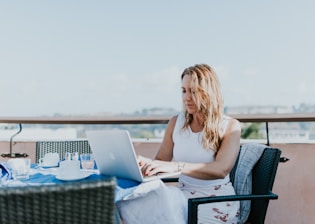A woman sits outdoors on a terrace, working on a laptop. She is dressed casually in a white top and patterned skirt. The table in front of her is covered with a blue tablecloth and holds several items, including a teacup and glasses. The background features a clear sky and blurred urban landscape.