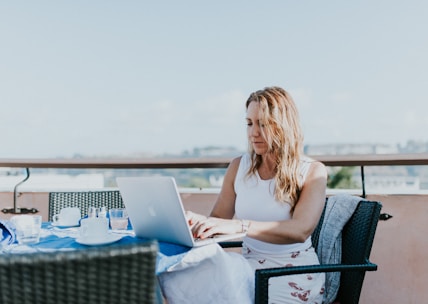 A woman sits outdoors on a terrace, working on a laptop. She is dressed casually in a white top and patterned skirt. The table in front of her is covered with a blue tablecloth and holds several items, including a teacup and glasses. The background features a clear sky and blurred urban landscape.