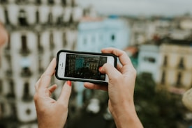 Hands holding a smartphone capturing a photograph of a cityscape, with colorful buildings visible in the background. The phone screen shows a focused image of the scene.