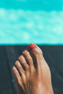 A foot with toenails painted in a bright color is in focus against a blurred background. The background features shades of turquoise, suggesting water, possibly from a pool.