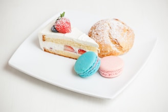 A white plate hosts an assortment of desserts, including a slice of cake with layers of cream and visible strawberry pieces, topped with a strawberry and blueberry. A round pastry dusted with powdered sugar and two colorful macarons, one blue and one pink, complete the arrangement.