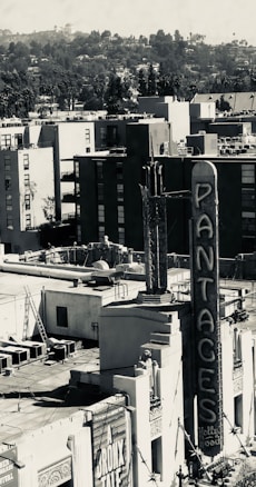 A grayscale urban scene with the iconic Pantages Theatre sign prominently featured in the foreground. Surrounding buildings in a dense city landscape are visible under a cloudy sky, with hills and trees in the distant background. The image presents a mix of architectural elements and city life.