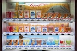 A vending machine stocked with various drinks, including soft drinks, juices, teas, and coffees. The machine displays colorful packaging and a range of brands like Coca-Cola, Fanta, and Georgia. Prices are listed in yen. A Japanese fan and traditional design elements decorate the background.