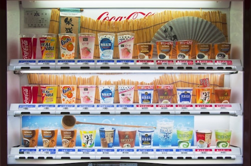 A vending machine stocked with various drinks, including soft drinks, juices, teas, and coffees. The machine displays colorful packaging and a range of brands like Coca-Cola, Fanta, and Georgia. Prices are listed in yen. A Japanese fan and traditional design elements decorate the background.