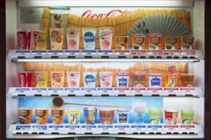 A vending machine stocked with various drinks, including soft drinks, juices, teas, and coffees. The machine displays colorful packaging and a range of brands like Coca-Cola, Fanta, and Georgia. Prices are listed in yen. A Japanese fan and traditional design elements decorate the background.