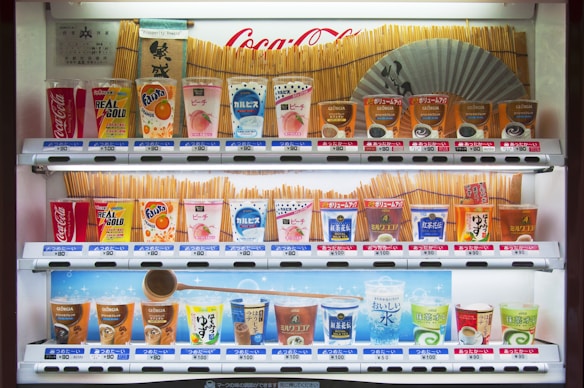 A vending machine stocked with various drinks, including soft drinks, juices, teas, and coffees. The machine displays colorful packaging and a range of brands like Coca-Cola, Fanta, and Georgia. Prices are listed in yen. A Japanese fan and traditional design elements decorate the background.