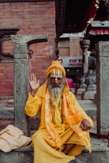 An elderly man with a long beard, dressed in bright yellow and orange robes, is sitting among stone pillars and raising his hand in a gesture. His face is adorned with traditional markings, and he wears large, round glasses. The background shows a brick wall and parts of a temple structure, with some signage visible in the distance.