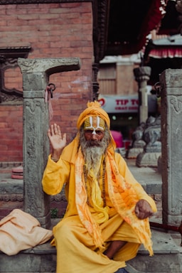 An elderly man with a long beard, dressed in bright yellow and orange robes, is sitting among stone pillars and raising his hand in a gesture. His face is adorned with traditional markings, and he wears large, round glasses. The background shows a brick wall and parts of a temple structure, with some signage visible in the distance.