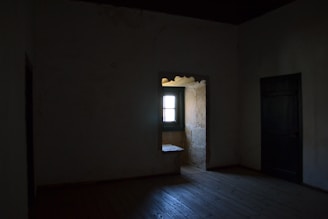 A dimly lit room with a single window casting gentle shadows on a wooden floor.