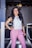 A woman is standing confidently in a corridor, wearing a stylish white sleeveless top and pink trousers. She has long, dark hair and is smiling, with one arm resting on the railing.