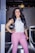 A woman is standing confidently in a corridor, wearing a stylish white sleeveless top and pink trousers. She has long, dark hair and is smiling, with one arm resting on the railing.