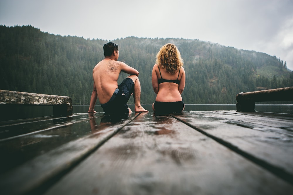 homem e mulher sentados na doca de madeira marrom do barco de frente para o corpo da água sob o céu cinzento