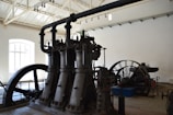 A large industrial machine with multiple vertical cylinders and pipes dominates the room. The setting appears to be an old factory or an industrial museum with high ceilings and a bright window letting in natural light. There is a large flywheel connected to the machinery, possibly part of a steam engine or similar type of mechanical equipment.