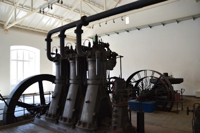 A large industrial machine with multiple vertical cylinders and pipes dominates the room. The setting appears to be an old factory or an industrial museum with high ceilings and a bright window letting in natural light. There is a large flywheel connected to the machinery, possibly part of a steam engine or similar type of mechanical equipment.