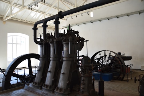 A large industrial machine with multiple vertical cylinders and pipes dominates the room. The setting appears to be an old factory or an industrial museum with high ceilings and a bright window letting in natural light. There is a large flywheel connected to the machinery, possibly part of a steam engine or similar type of mechanical equipment.