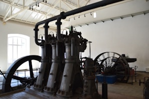 A large industrial machine with multiple vertical cylinders and pipes dominates the room. The setting appears to be an old factory or an industrial museum with high ceilings and a bright window letting in natural light. There is a large flywheel connected to the machinery, possibly part of a steam engine or similar type of mechanical equipment.