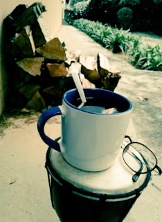 A white mug with a blue rim and handle sits on top of a drum, accompanied by a pair of eyeglasses. A spoon is inside the mug, which likely contains a dark liquid, possibly coffee or tea. In the background, there is a stack of cut logs against a wall and a pathway lined with green plants.