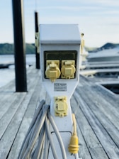 An electrical outlet station with multiple yellow power sockets is mounted on a wooden dock. Thick grey cables are coiled around the base of the station, with one yellow plug prominently hanging. In the background, calm water and blurry outlines of hills can be observed. The overall setting suggests a marina or docking area.