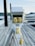 An electrical outlet station with multiple yellow power sockets is mounted on a wooden dock. Thick grey cables are coiled around the base of the station, with one yellow plug prominently hanging. In the background, calm water and blurry outlines of hills can be observed. The overall setting suggests a marina or docking area.