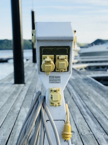 An electrical outlet station with multiple yellow power sockets is mounted on a wooden dock. Thick grey cables are coiled around the base of the station, with one yellow plug prominently hanging. In the background, calm water and blurry outlines of hills can be observed. The overall setting suggests a marina or docking area.