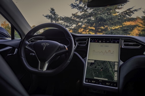 An interior view of a car focusing on the steering wheel and a large central touchscreen displaying a map and a graph. The car is parked outdoors with a view of trees through the windshield.