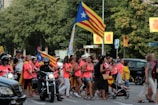 A group of people marching on a street, holding flags characterized by red and yellow stripes with a white star on a blue triangle. Many participants wearing bright pink tops, walking alongside vehicles including a motorcycle and a car. Green trees and various street signs are visible in the background.