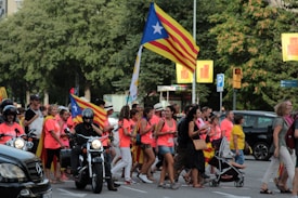 A group of people marching on a street, holding flags characterized by red and yellow stripes with a white star on a blue triangle. Many participants wearing bright pink tops, walking alongside vehicles including a motorcycle and a car. Green trees and various street signs are visible in the background.