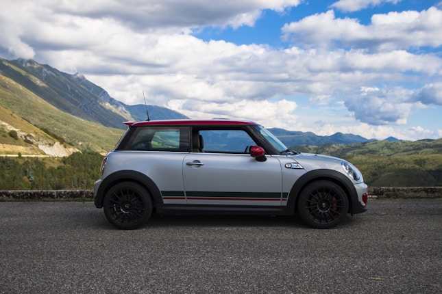 A silver and red compact car parked on an asphalt road with a scenic mountain landscape in the background. The sky is partly cloudy, adding contrast to the green hills and distant peaks.