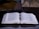 An open book with dense text lies on a dark, glossy surface, accompanied by a pen resting horizontally between the pages. In the background, a laptop with a partially visible screen enhances the academic atmosphere.