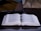 An open book with dense text lies on a dark, glossy surface, accompanied by a pen resting horizontally between the pages. In the background, a laptop with a partially visible screen enhances the academic atmosphere.