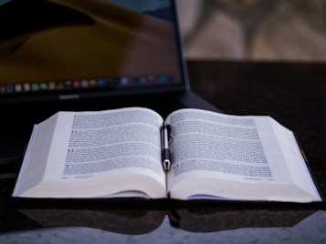 An open book with dense text lies on a dark, glossy surface, accompanied by a pen resting horizontally between the pages. In the background, a laptop with a partially visible screen enhances the academic atmosphere.