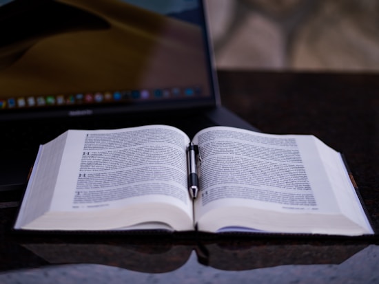 An open book with dense text lies on a dark, glossy surface, accompanied by a pen resting horizontally between the pages. In the background, a laptop with a partially visible screen enhances the academic atmosphere.