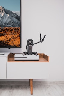 On a light wooden media console, a small drone with propellers is placed in front of a flat-screen TV. The TV displays a landscape image with snow-capped mountains and a field with golden vegetation. The console also holds a gaming console beneath the drone.
