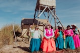 A group of women wearing vibrant, colorful traditional clothing stand together on a grassy, sunlit area with woven structures in the background. Their attire includes bright skirts, embroidered tops, and woven hats.