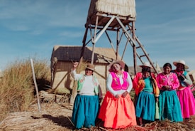 A group of women wearing vibrant, colorful traditional clothing stand together on a grassy, sunlit area with woven structures in the background. Their attire includes bright skirts, embroidered tops, and woven hats.
