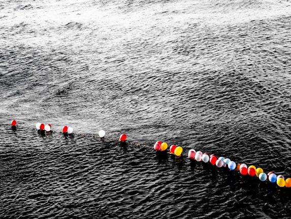A line of colorful balloons floats on the surface of dark, wavy water. The balloons are red, white, yellow, and some mixed with blue. The water appears deep and slightly turbulent, contrasting with the vibrant colors of the balloons.
