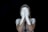 A person with light skin is standing against a black background, covering their face with hands that are painted white, extending the white paint to parts of their arms and shoulders.