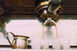 Espresso coffee is pouring from a machine into two white ceramic cups, with a metal milk frother pitcher placed nearby.