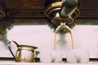 Espresso coffee is pouring from a machine into two white ceramic cups, with a metal milk frother pitcher placed nearby.
