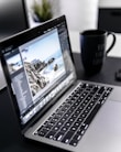 A laptop displaying photo editing software is situated on a dark desk with a large black coffee mug nearby. The screen shows an image of a coastal landscape with rocks and ocean.