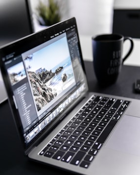 A laptop displaying photo editing software is situated on a dark desk with a large black coffee mug nearby. The screen shows an image of a coastal landscape with rocks and ocean.