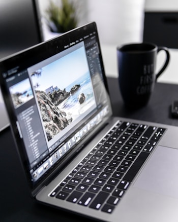 A laptop displaying photo editing software is situated on a dark desk with a large black coffee mug nearby. The screen shows an image of a coastal landscape with rocks and ocean.