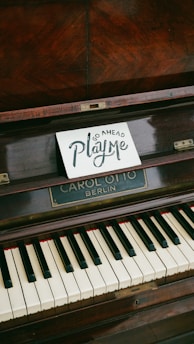 A wooden upright piano displays a small sign on its music stand, inviting interaction with the words 'Go Ahead Play Me' in artistic lettering. The piano has visible keys and a nameplate reading 'CAROL OTTO BERLIN'.