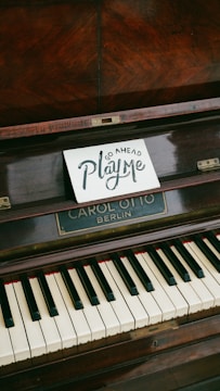 A wooden upright piano displays a small sign on its music stand, inviting interaction with the words 'Go Ahead Play Me' in artistic lettering. The piano has visible keys and a nameplate reading 'CAROL OTTO BERLIN'.