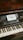 A wooden upright piano displays a small sign on its music stand, inviting interaction with the words 'Go Ahead Play Me' in artistic lettering. The piano has visible keys and a nameplate reading 'CAROL OTTO BERLIN'.