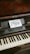 A wooden upright piano displays a small sign on its music stand, inviting interaction with the words 'Go Ahead Play Me' in artistic lettering. The piano has visible keys and a nameplate reading 'CAROL OTTO BERLIN'.