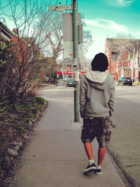 A person is walking down a quiet urban street next to a sidewalk lined with bare trees and bushes. They wear a gray hoodie, plaid shorts, and black sneakers. The street sign indicates Ellsworth Ave, and several buildings and parked cars are visible along the street. The sky above is clear with some clouds.
