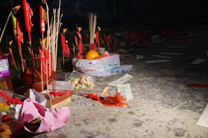 A collection of incense sticks and red candles is set up outdoors, possibly for a ritual or offering. Papers with text, some partially burnt, and various objects including fruits and packaged items are placed on the ground. The setting is dimly lit, providing a solemn and reflective atmosphere.