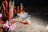 A collection of incense sticks and red candles is set up outdoors, possibly for a ritual or offering. Papers with text, some partially burnt, and various objects including fruits and packaged items are placed on the ground. The setting is dimly lit, providing a solemn and reflective atmosphere.